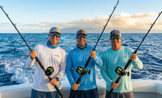 Three smiling anglers on a moving sport fishing boat hold up their Eat My Tackle deep sea rods, with a wide blue ocean and distant sunlit clouds in the background.